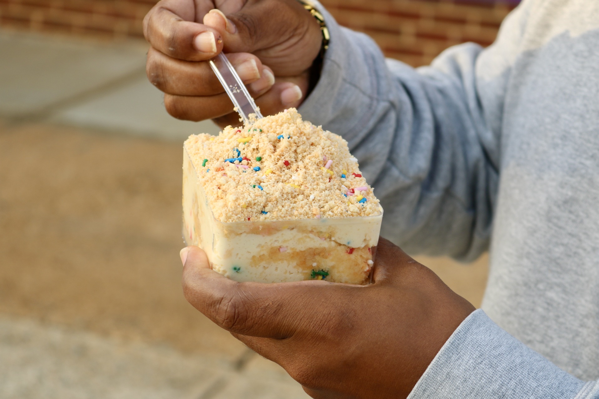 Person holding a slice of cake with colorful sprinkles, using a fork outdoors, wearing a gray sweatshirt.