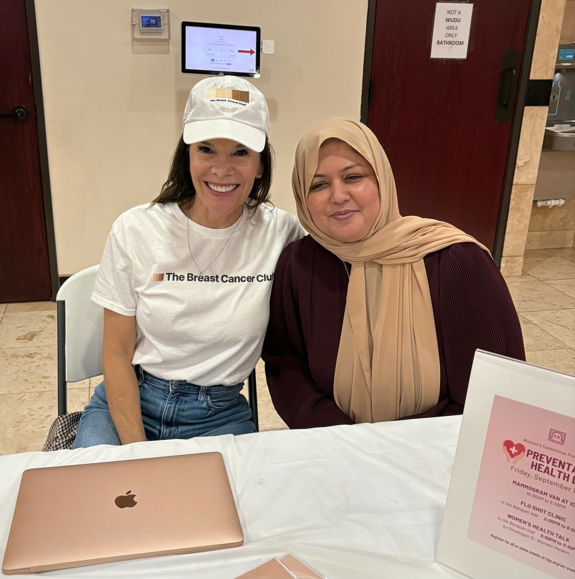 Two women sitting at a table, smiling, with a laptop and a framed sign in front of them.