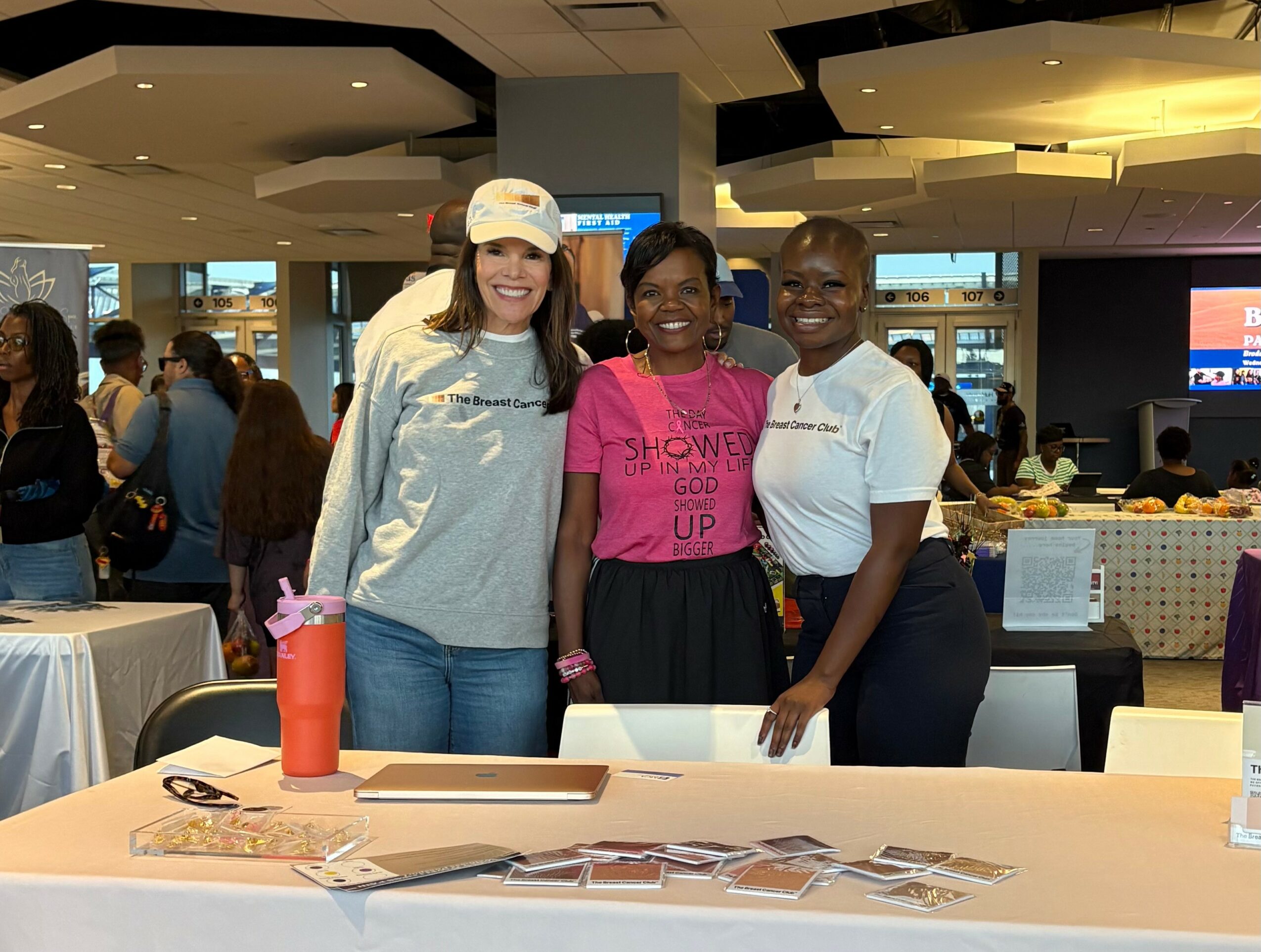 Three women standing together indoors, smiling, with tables and people in the background.