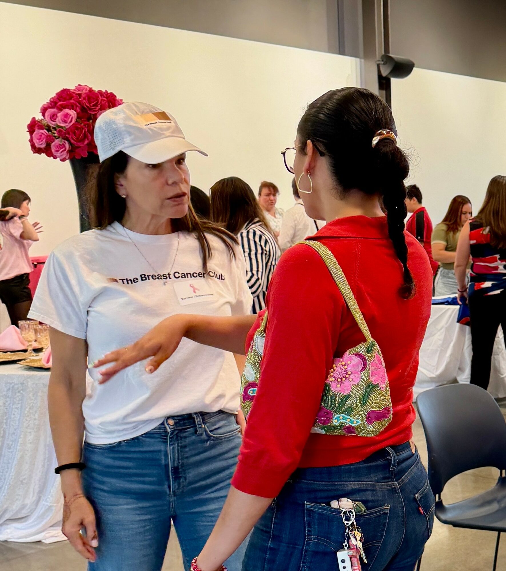 Two women are talking indoors, one wearing a white t-shirt and cap, the other in a red sweater with a floral bag.