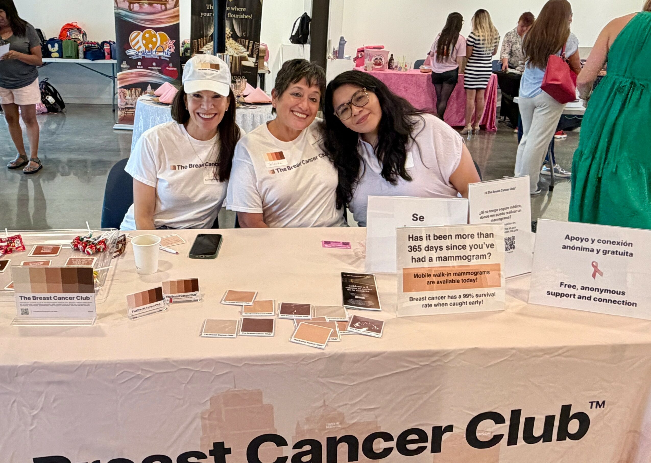 Three women sit behind a table at a breast cancer awareness event, smiling, with informational materials displayed.