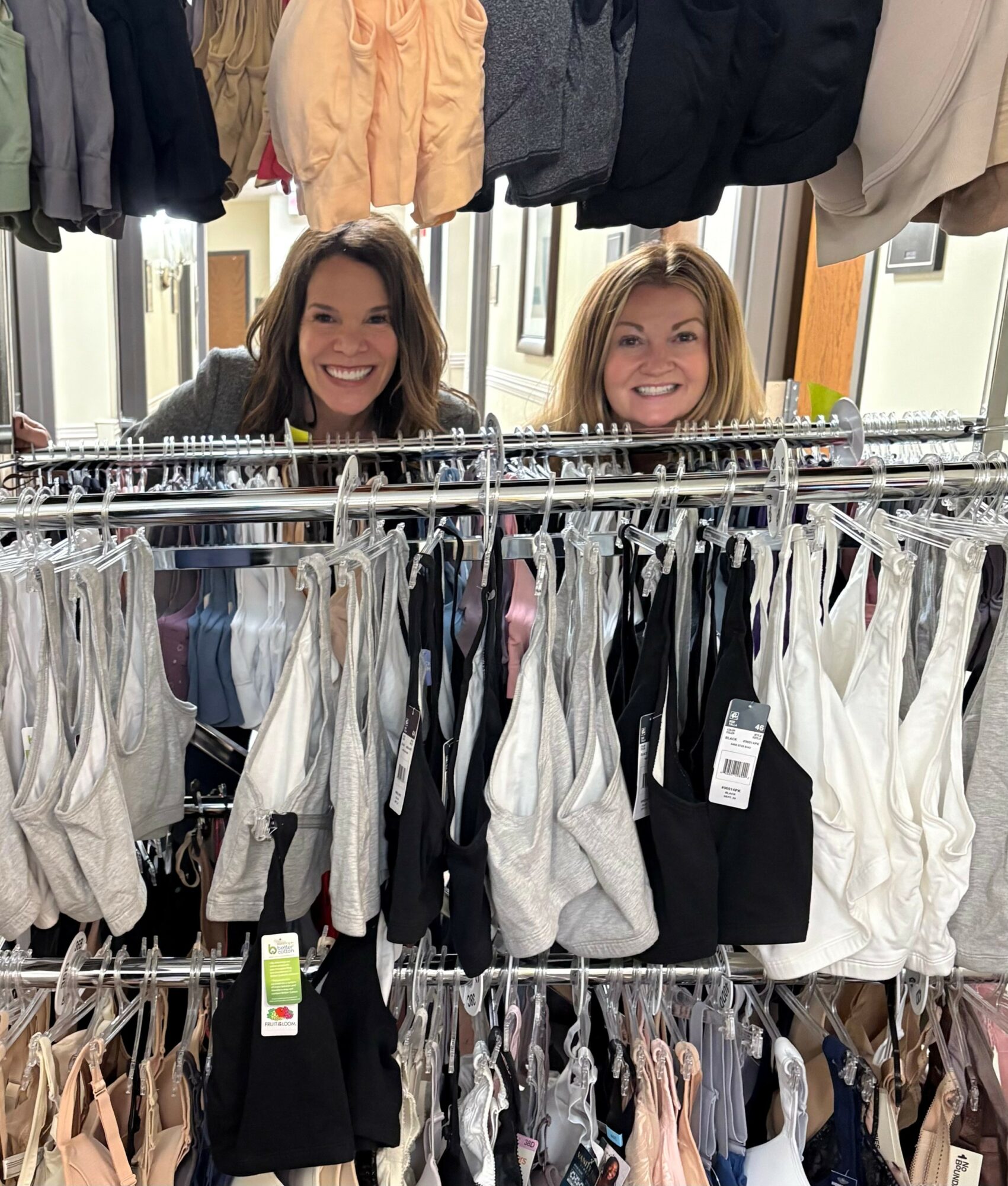 Two women smiling behind clothing racks with various garments, in a store or boutique setting.