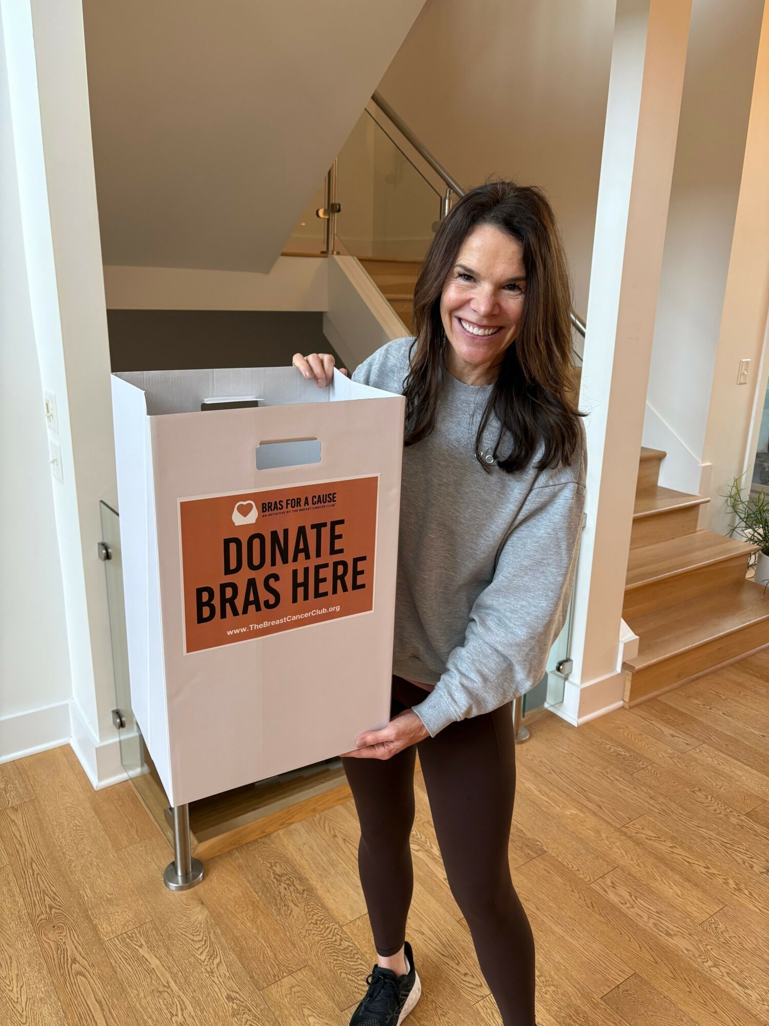 Woman smiling, standing next to a donation bin labeled 'Donate Brakes Here,' indoors with wooden flooring.