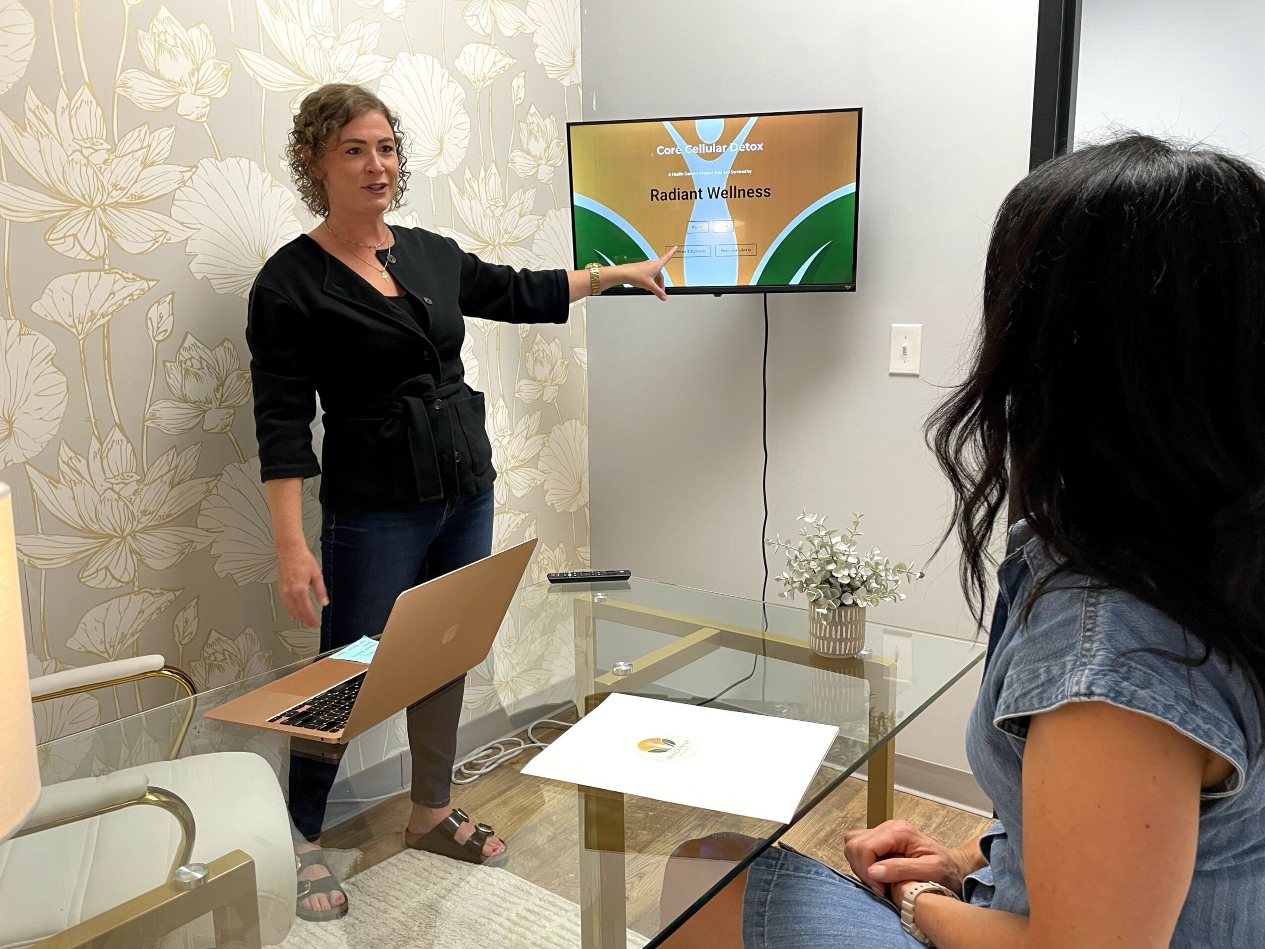 Woman presenting to a seated woman in a meeting room with a wall-mounted screen, laptop, and paper on the table.