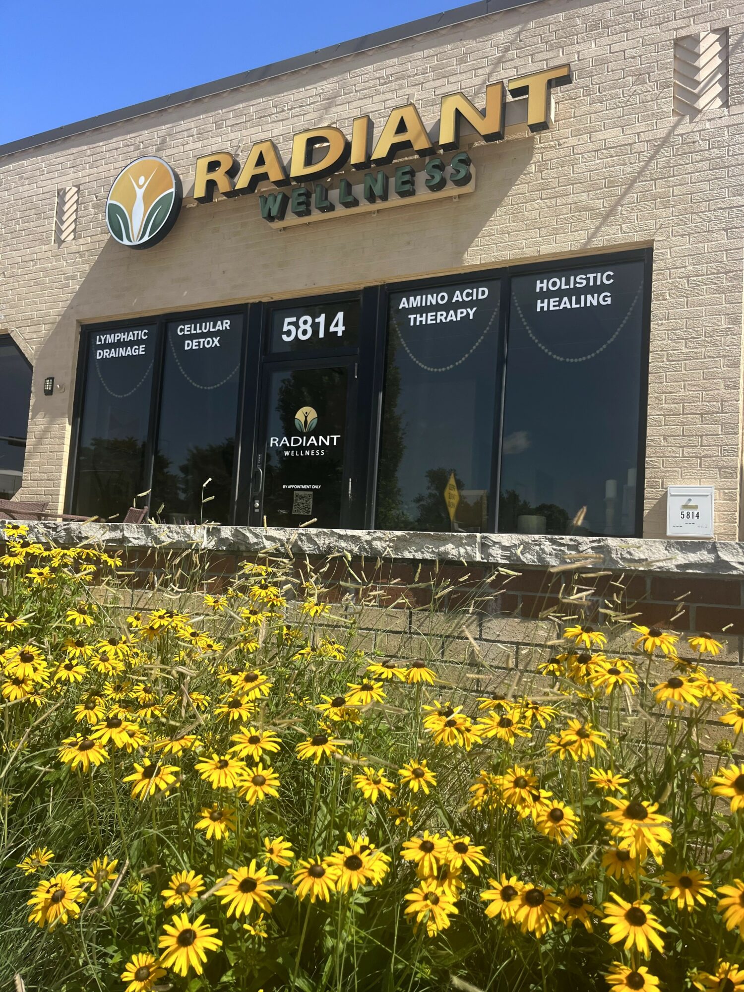 Storefront with sign reading 'RADIANT' and yellow flowers in front, blue sky background.