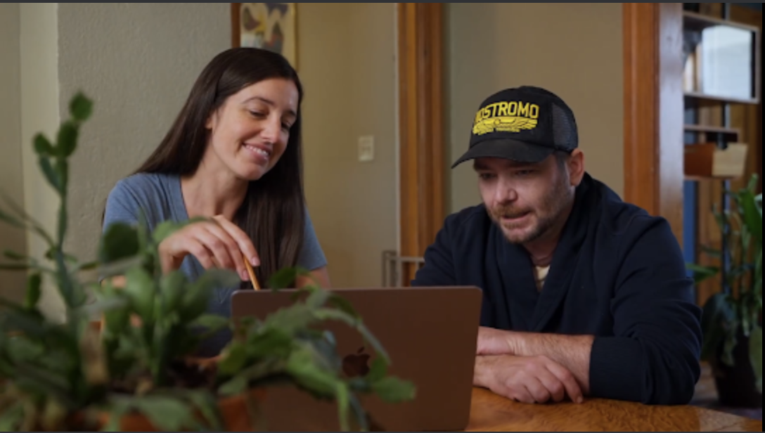 Two people sit at a table looking at a laptop, smiling, in a room with plants and wooden furniture.