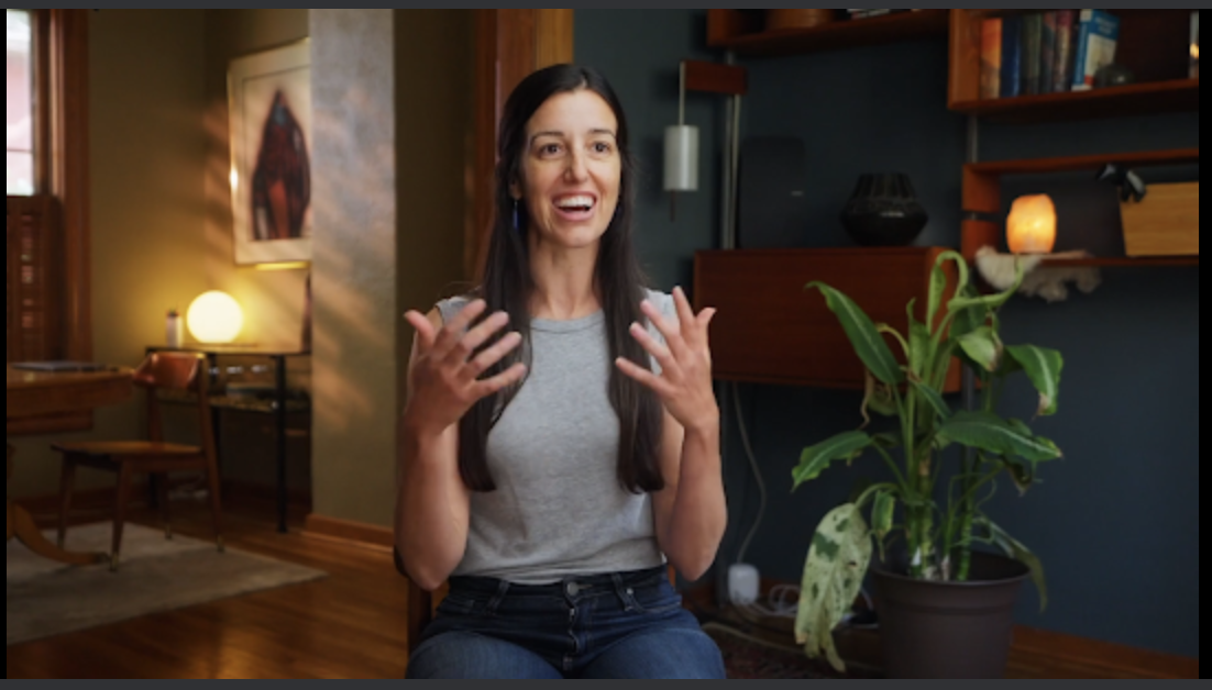 Woman sitting indoors, gesturing with hands, smiling, in a room with books and plants.