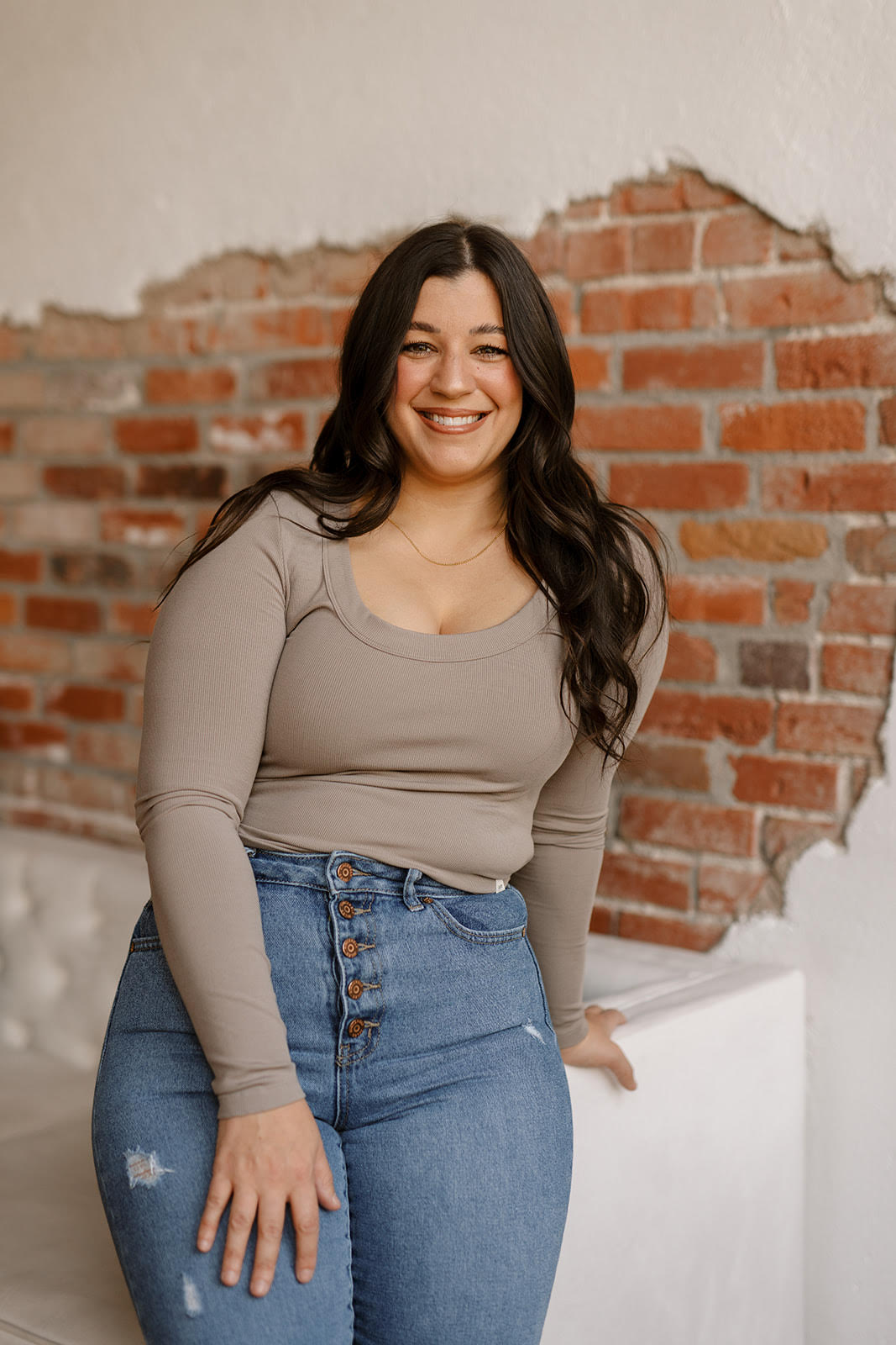 Smiling woman with long dark hair in a beige top and jeans, standing against a brick wall.