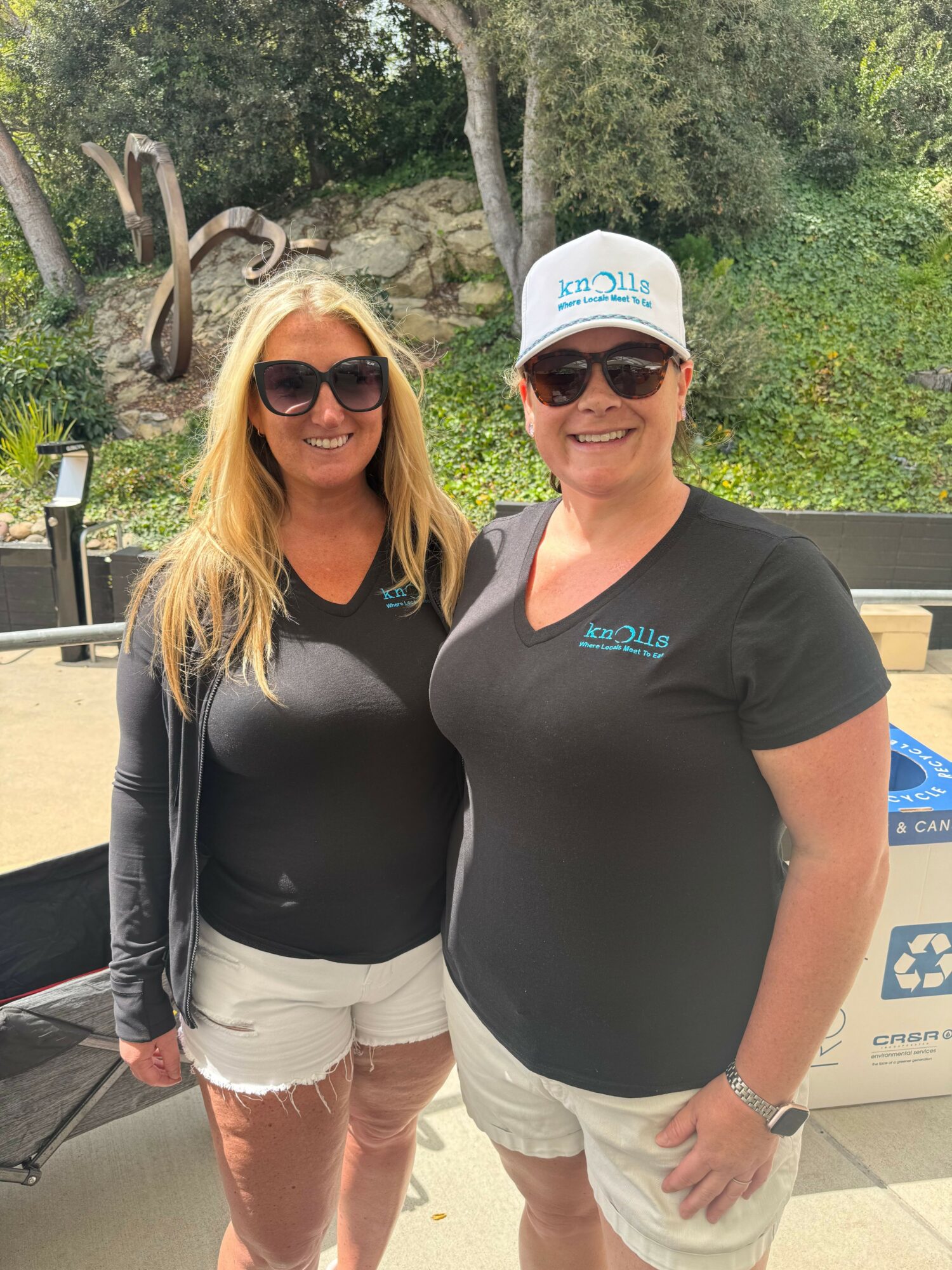 Two women standing outdoors, smiling, wearing sunglasses and casual clothing, with trees and rocks in the background.