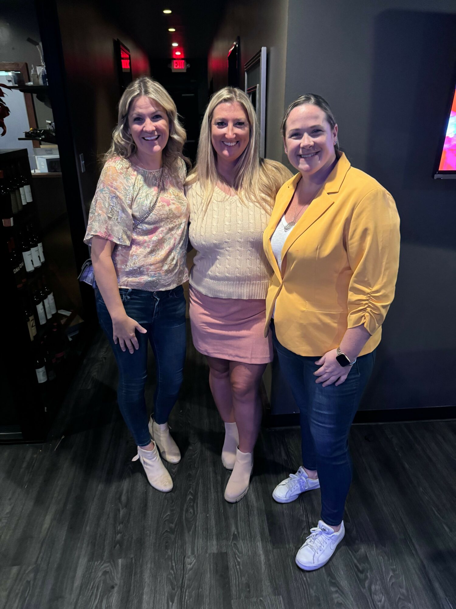 Three women standing together indoors, smiling, with dark flooring and a dark wall in the background.