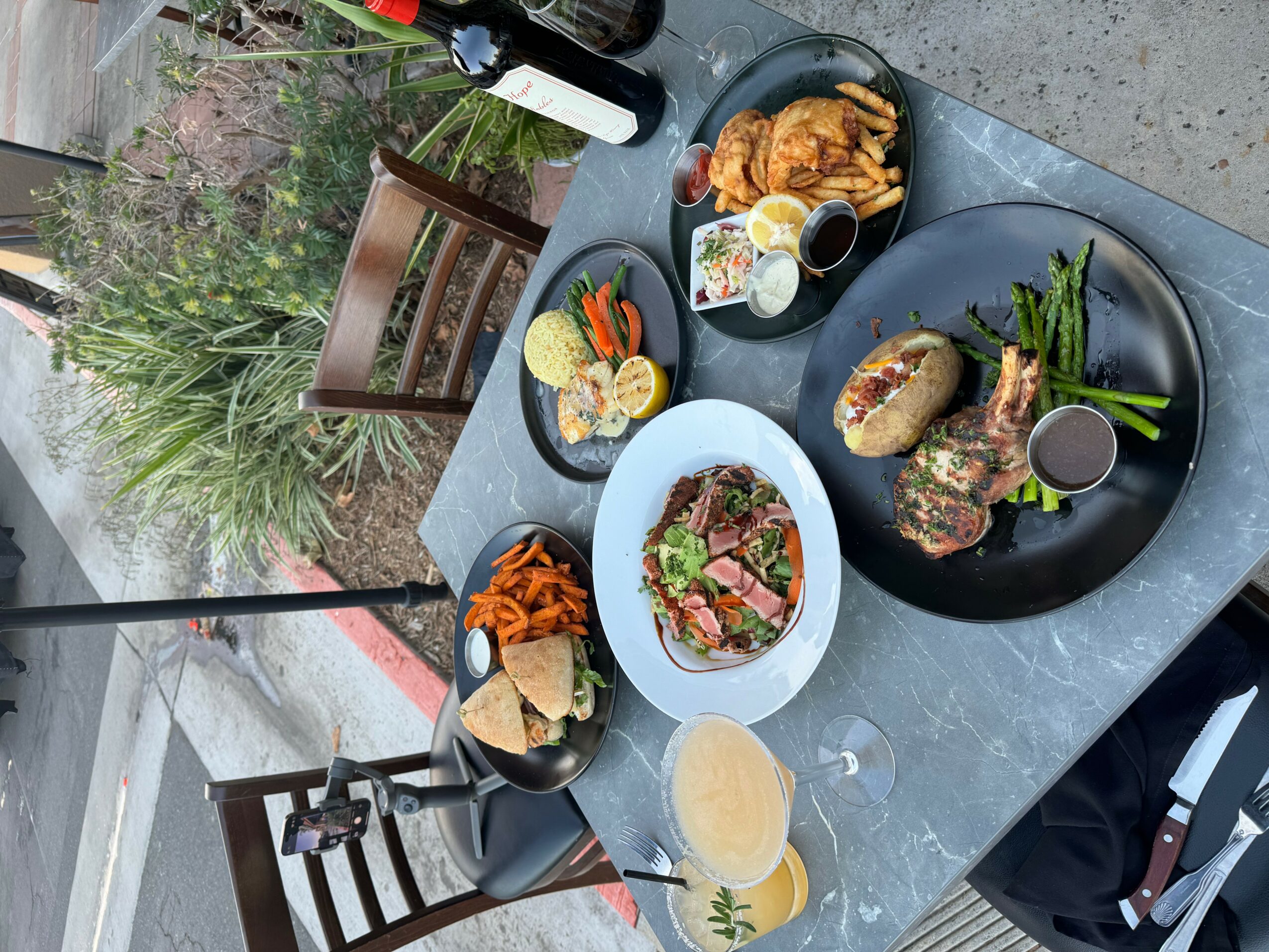 Outdoor table with various dishes including salad, grilled meat, vegetables, and drinks, with a garden background.