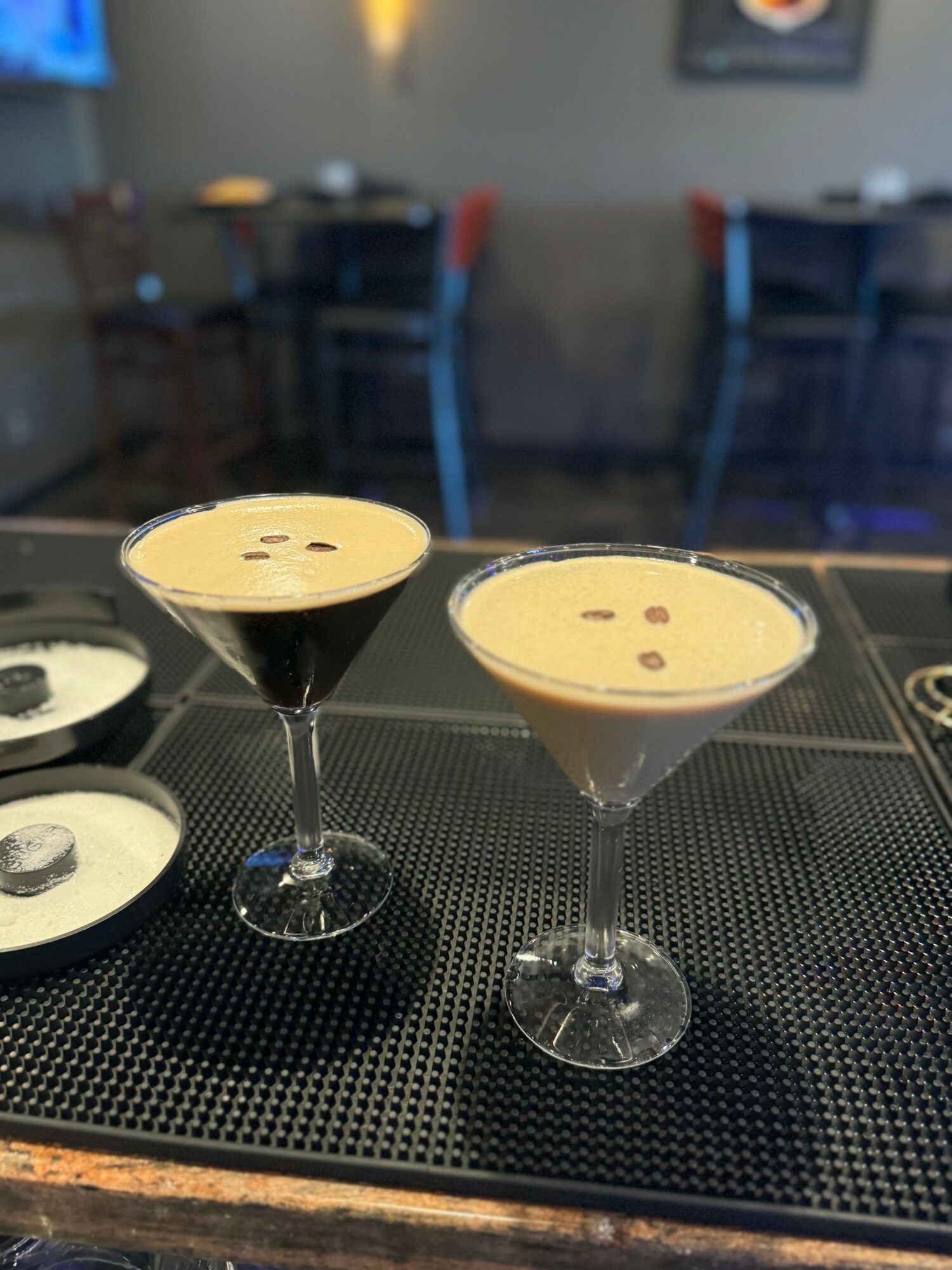 Two cocktails in martini glasses on a bar counter with a blurred background of chairs and tables.
