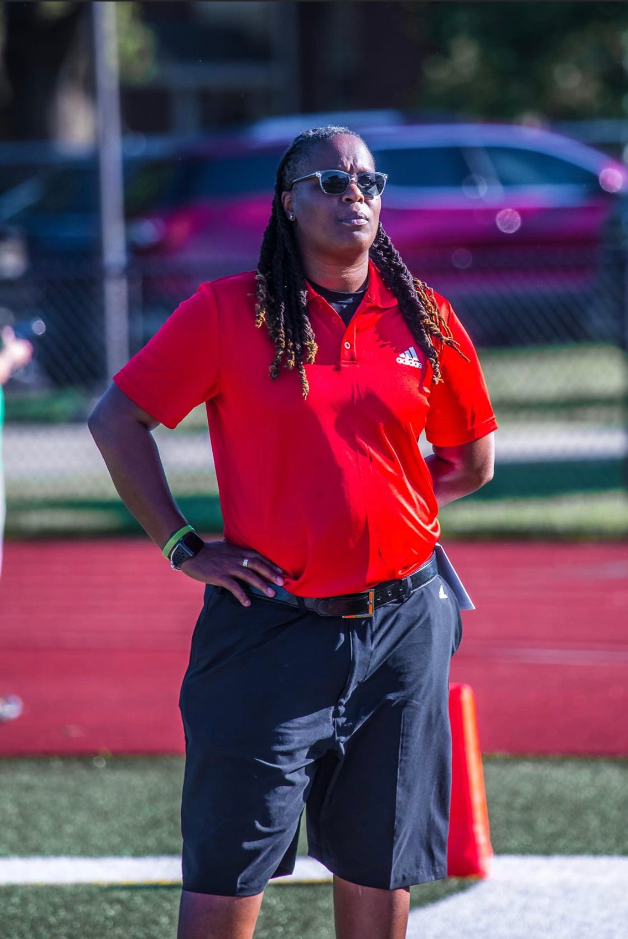 Woman in red shirt and black shorts standing outdoors with hands on hips, wearing sunglasses, near a tennis court.