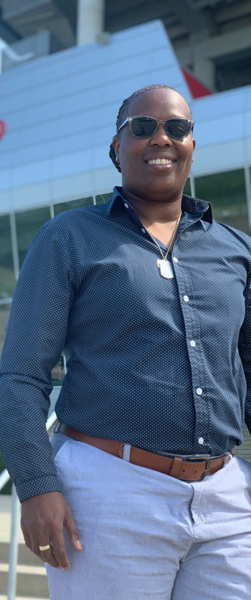 Woman wearing sunglasses, dark blue shirt, light pants, standing outdoors in front of a modern building.