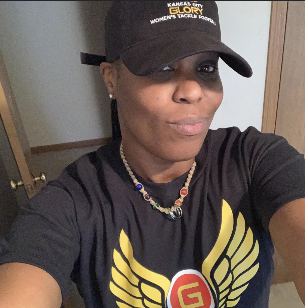 Woman wearing a black cap and T-shirt, smiling, with a necklace, indoors, in front of a door.