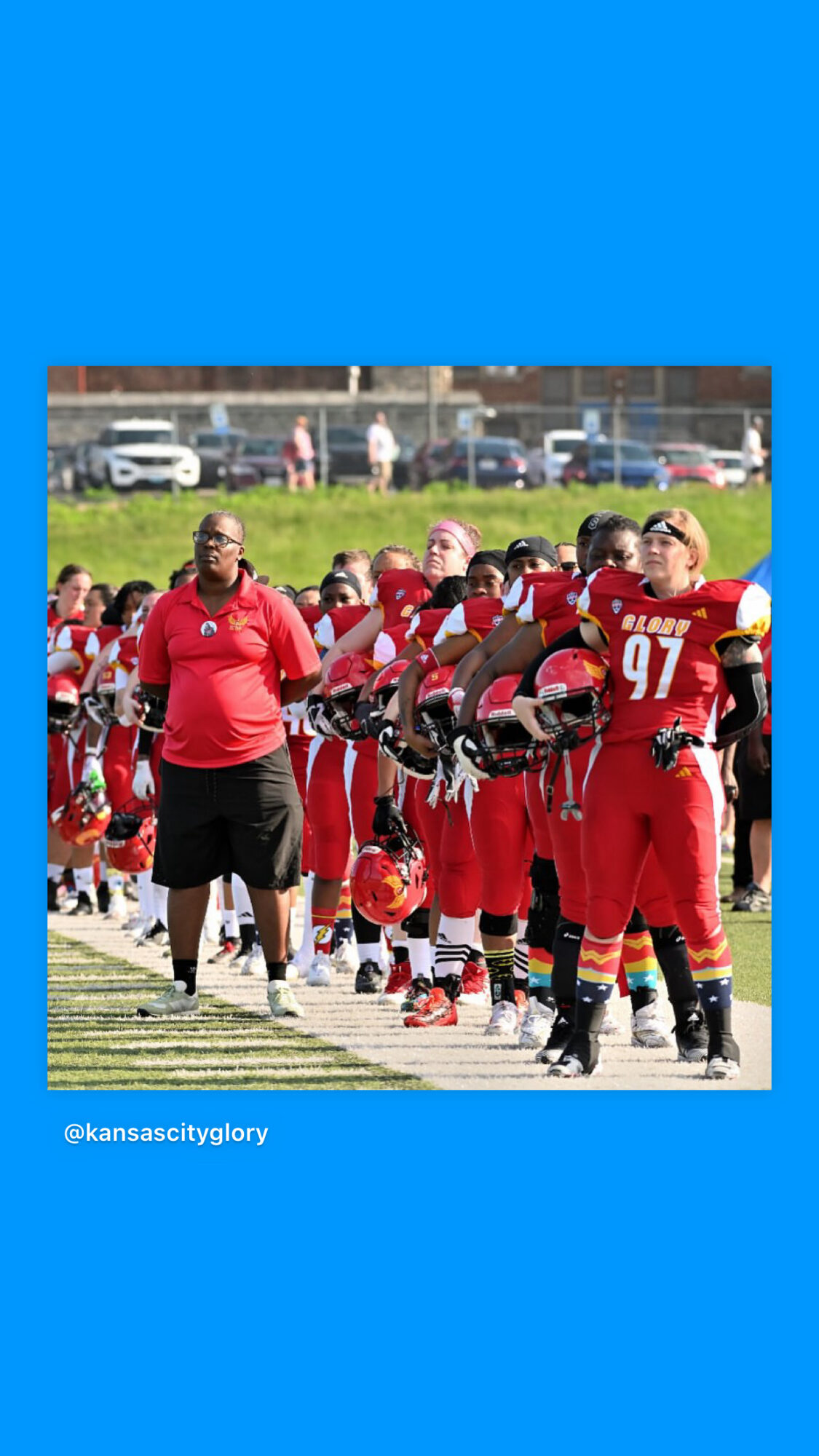 Football team in red uniforms standing on field, coach nearby, spectators and cars in background, blue border.