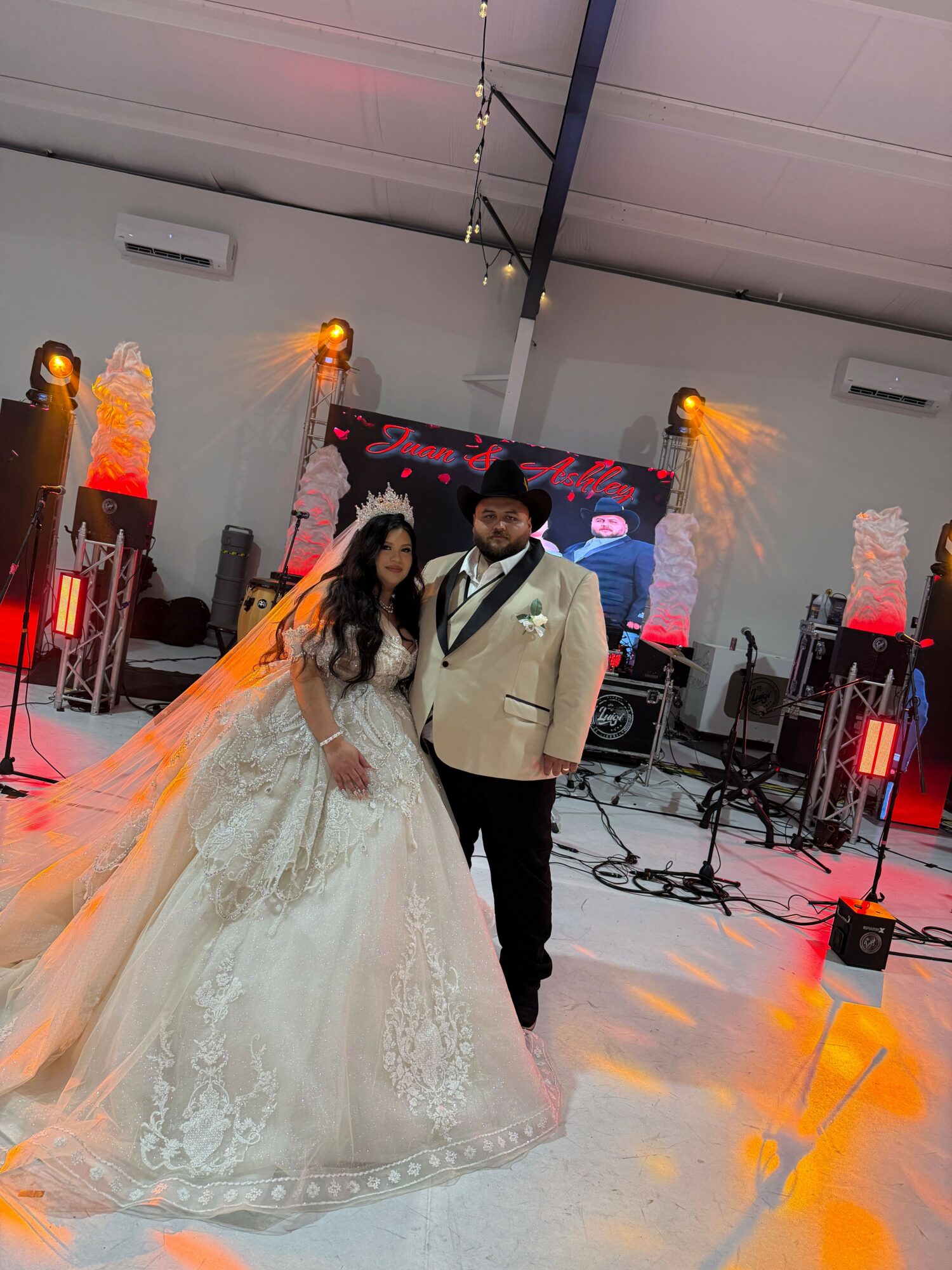 A couple dancing, the woman in a white gown and the man in a beige jacket, in a decorated event space with stage lighting.