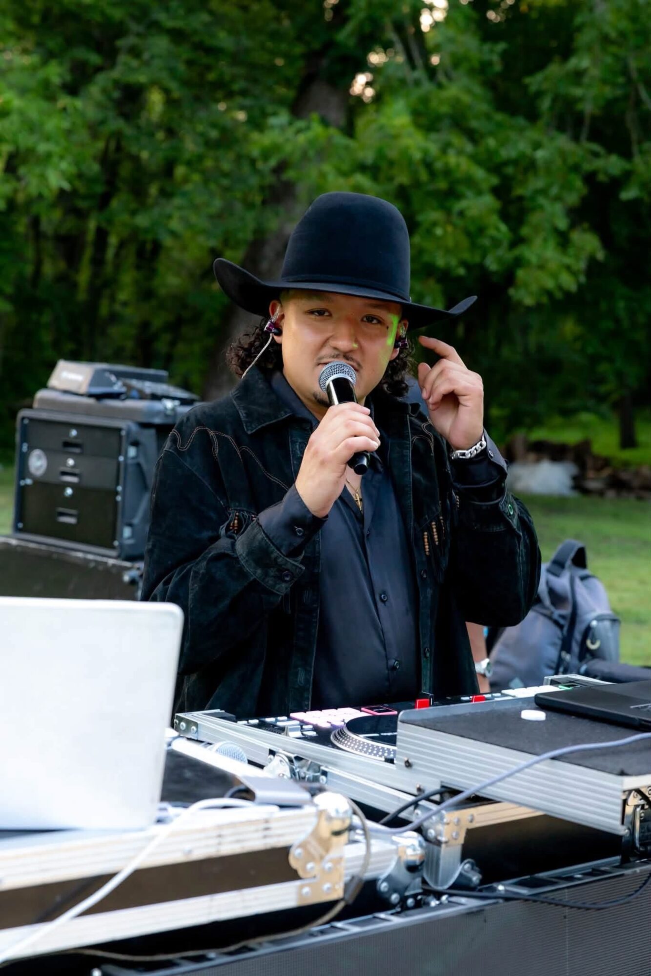 Woman wearing a black hat and jacket speaking into a microphone outdoors with trees in background.