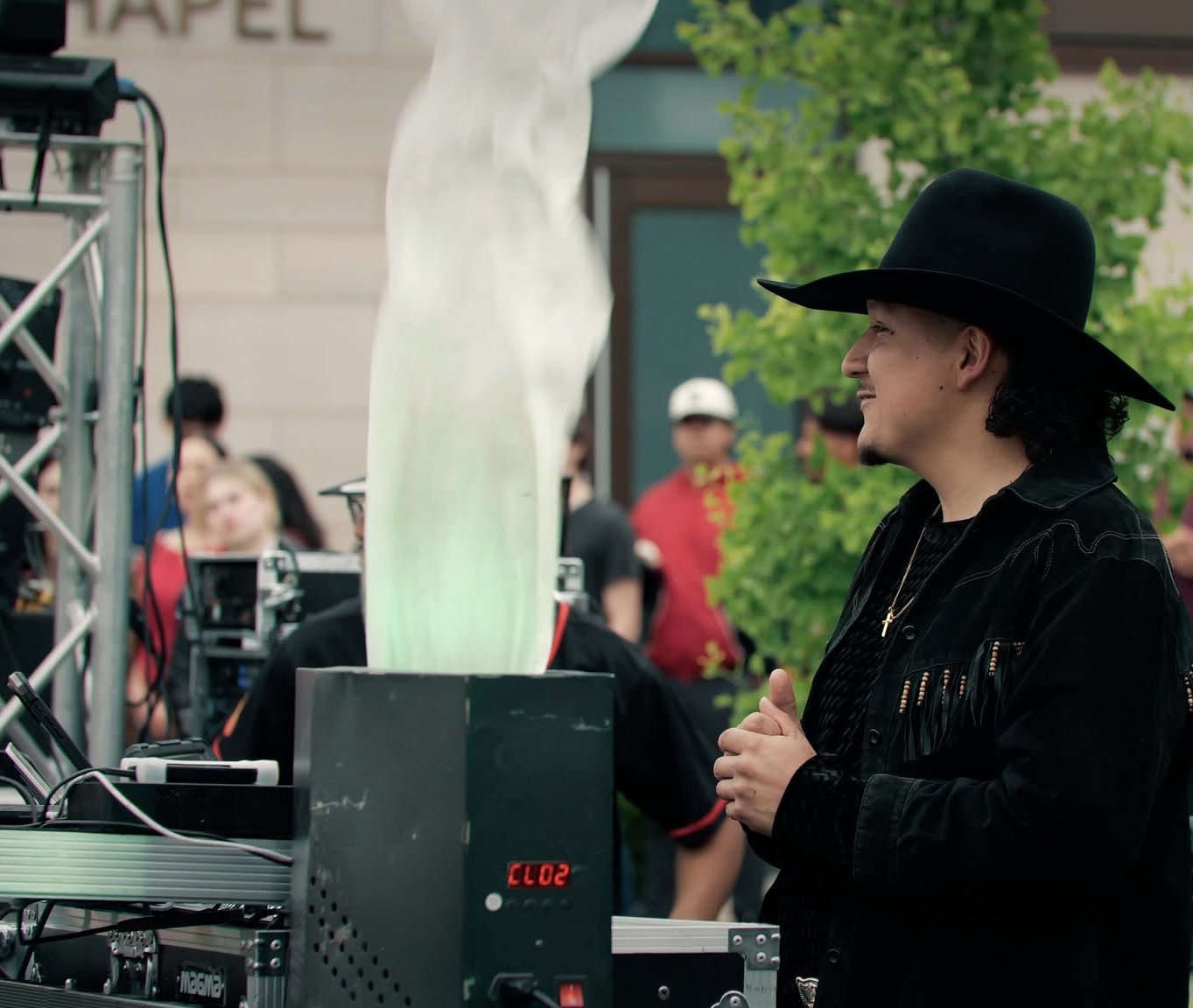 Person wearing a black hat and jacket stands near a device emitting green smoke, with people and trees in background.