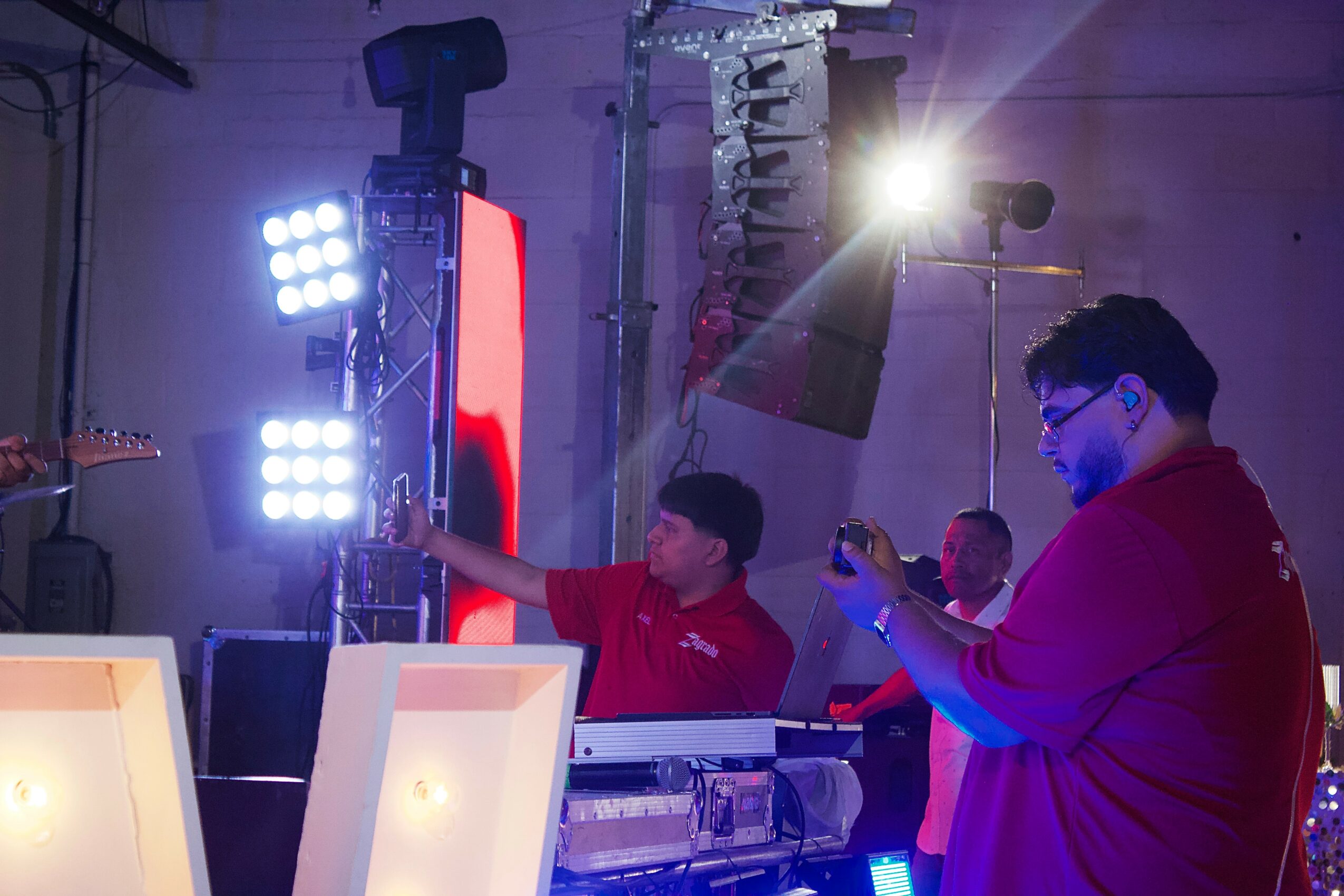 People in red shirts working with lighting and equipment in a studio or stage setting, bright lights overhead.