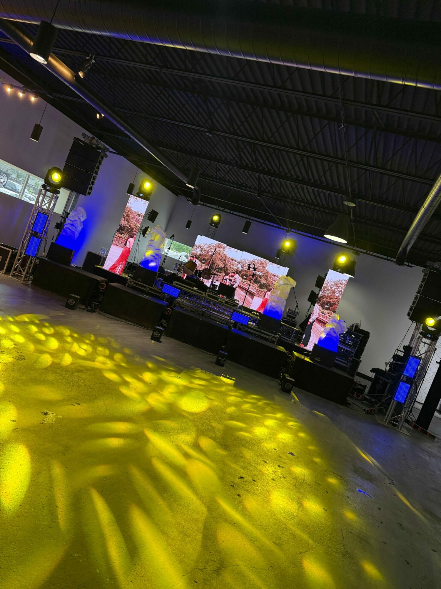 Indoor event space with a stage, large screens, and yellow lighting on the floor, viewed from the side.