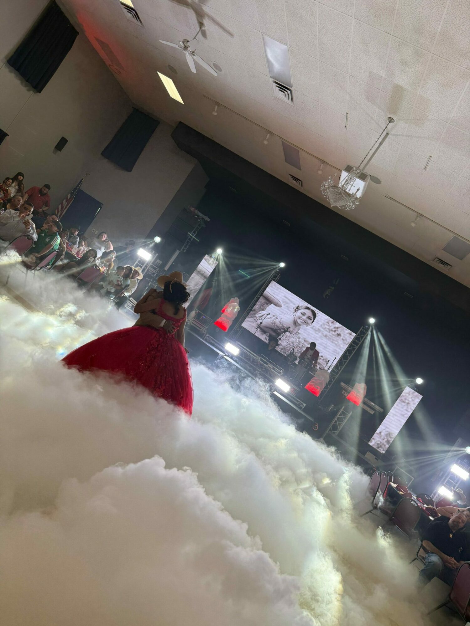 Person in red coat standing in front of stage with bright lights and screens, surrounded by fog or smoke.