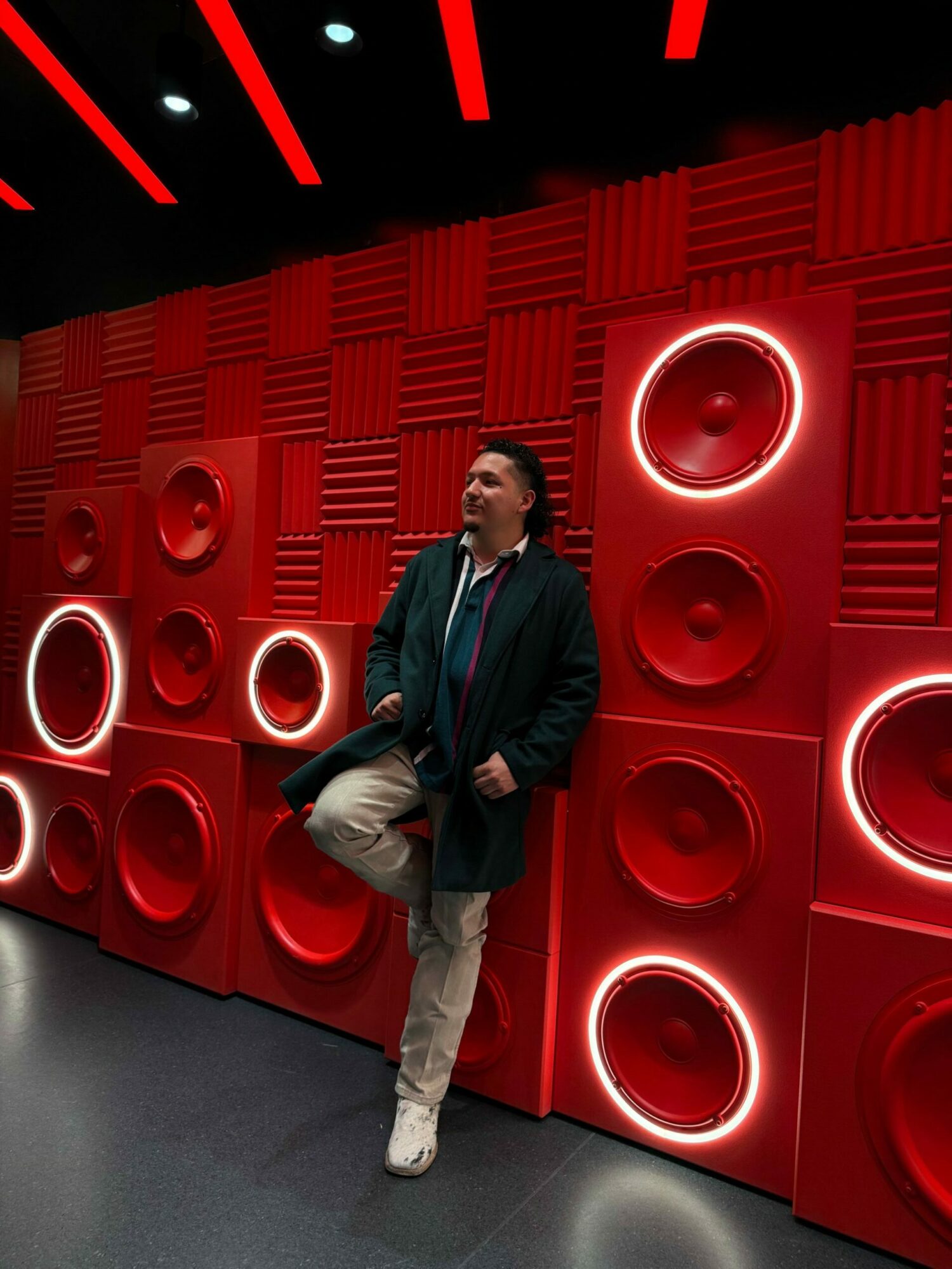 Person sitting against a wall of red speakers with circular lights, in a dark room.