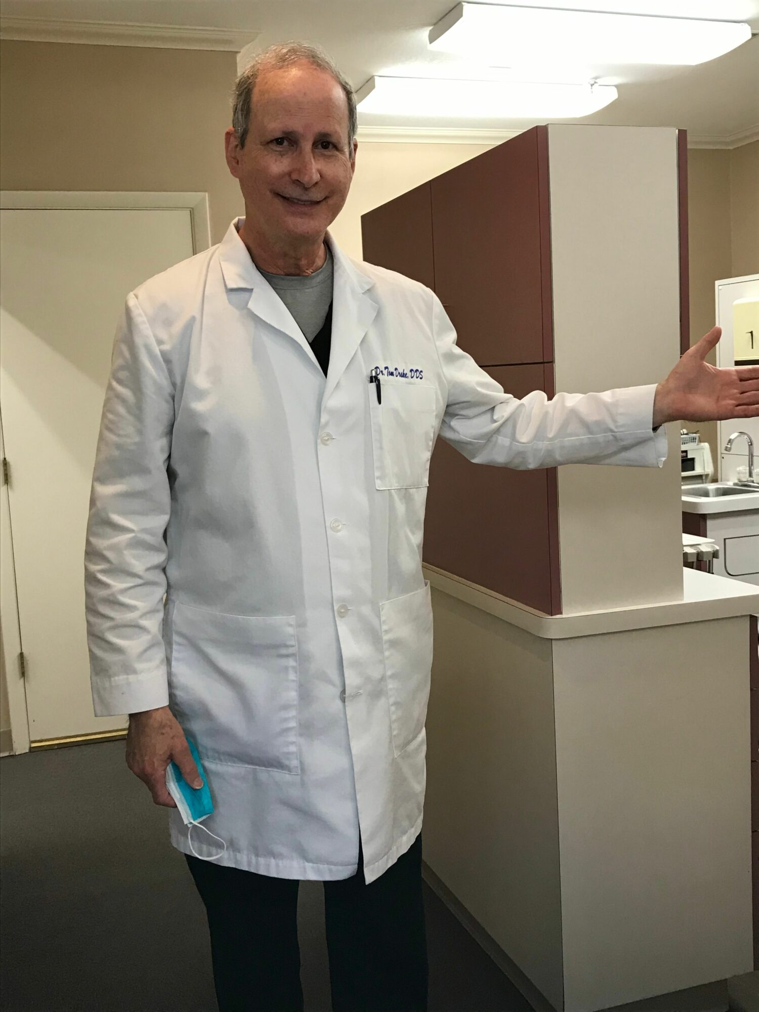 Person in a white lab coat standing in a room, waving with one hand, holding a blue object in the other.