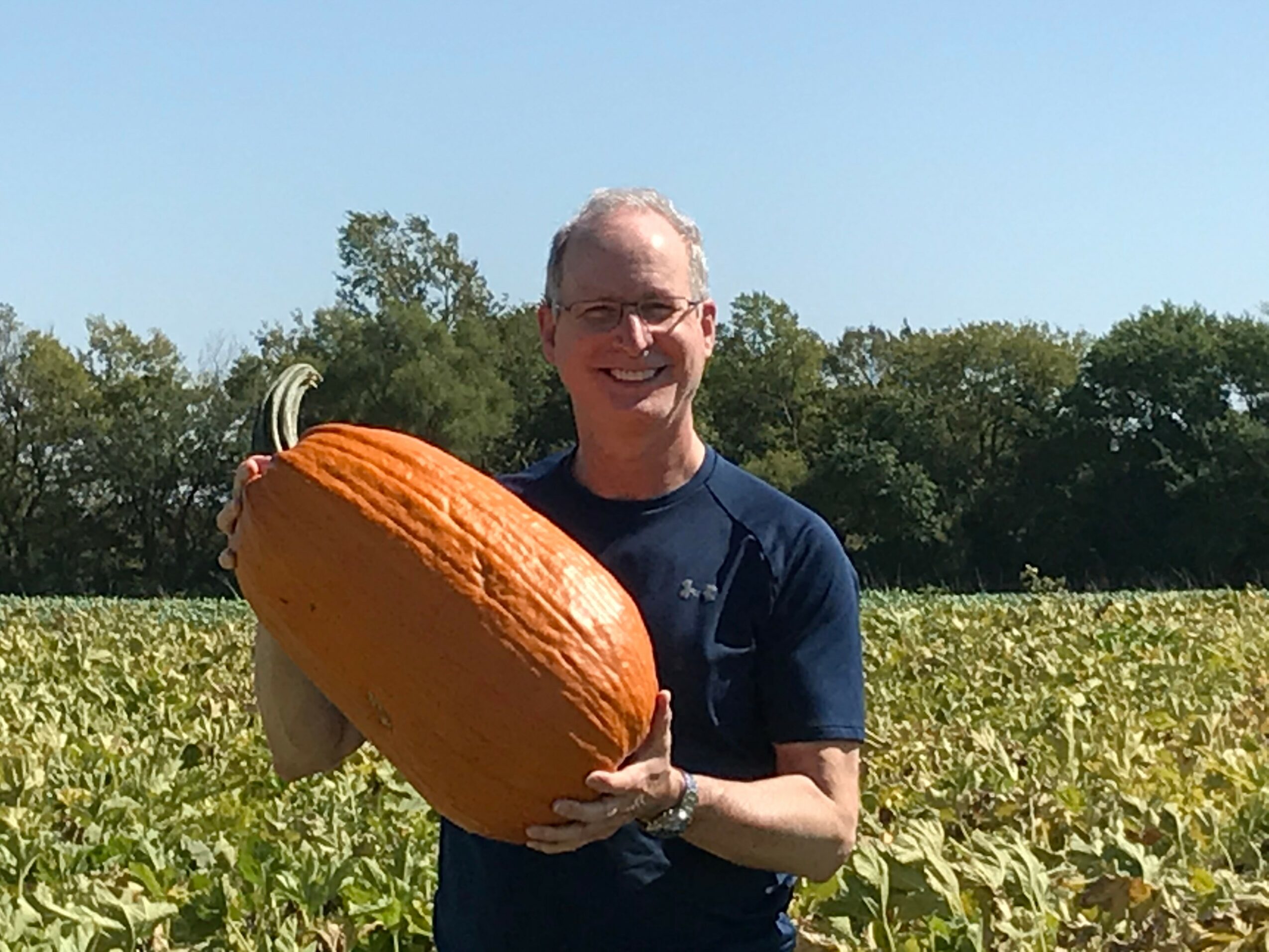 Man holding large pumpkin in a field with trees in the background, smiling, sunny day.