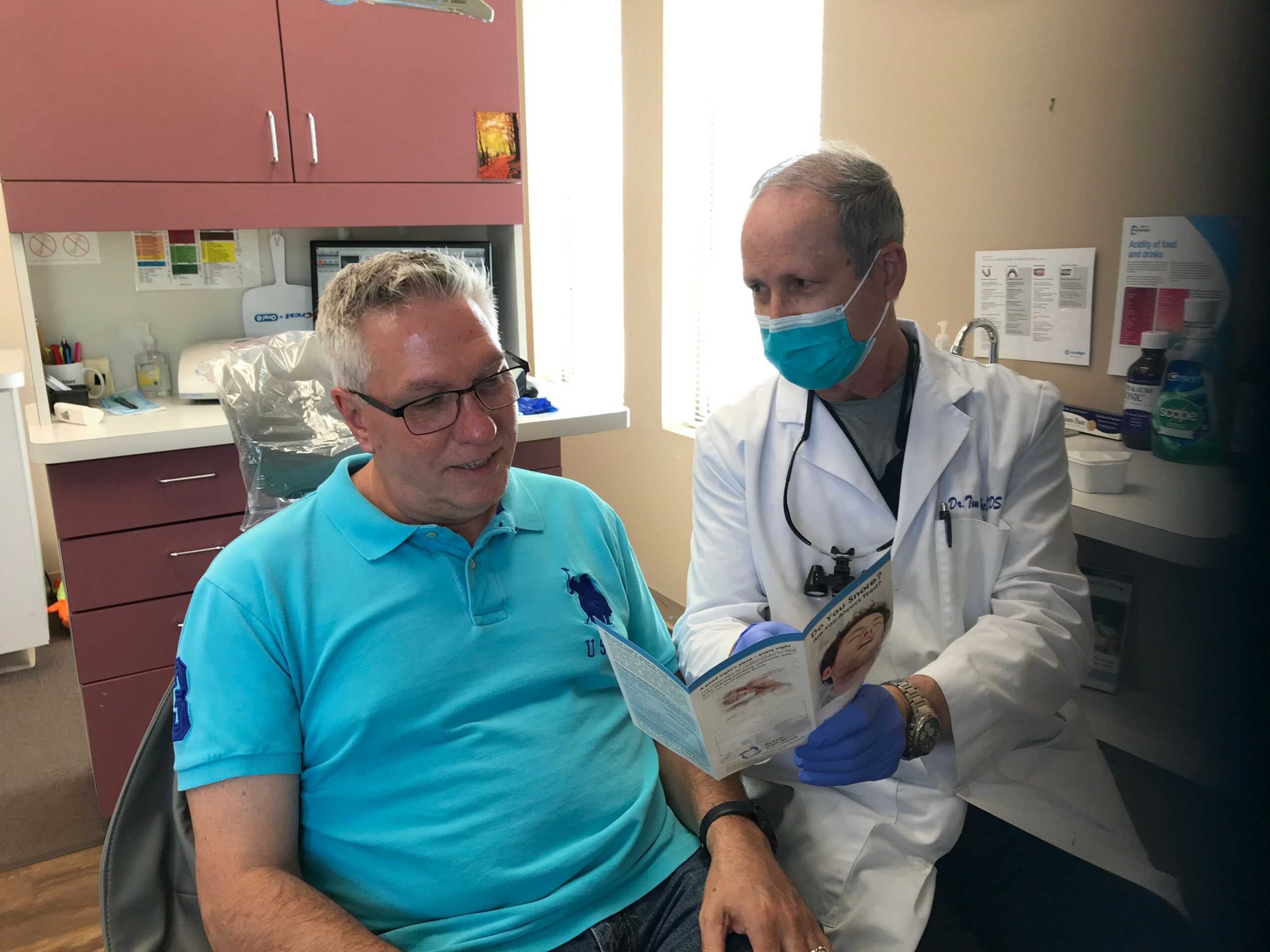 Doctor and patient in a medical office, doctor showing a booklet to the patient, both wearing masks, patient in a blue shirt.