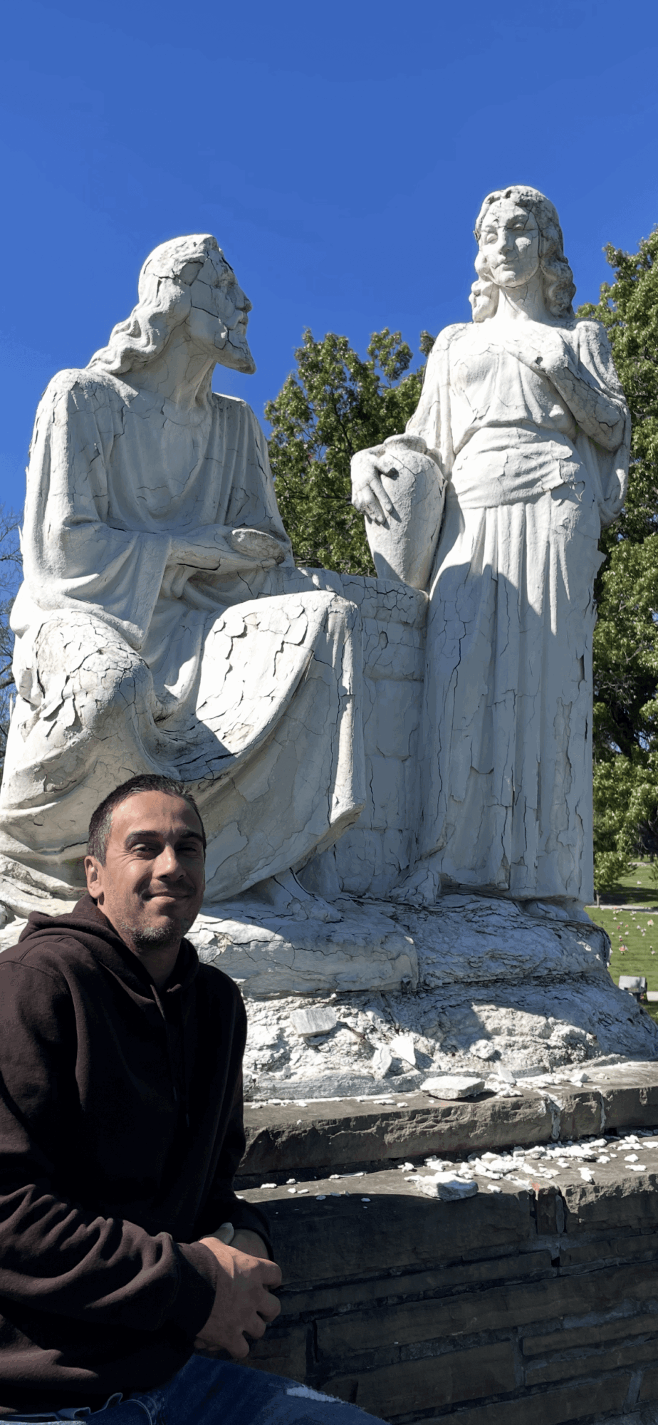 Person sitting in front of a large white statue of two women, with trees and blue sky in background.