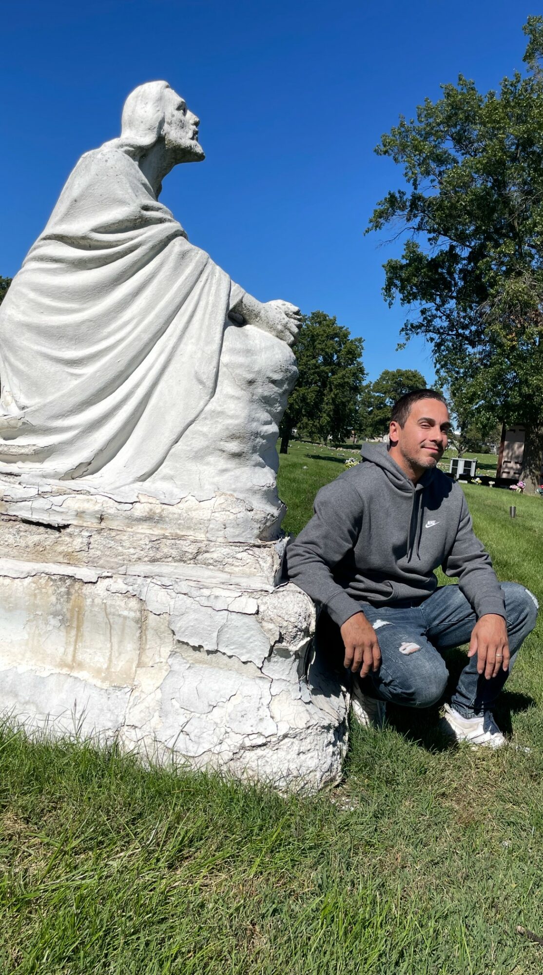Young man crouching next to a large white statue of a seated figure outdoors, with trees and blue sky in background.