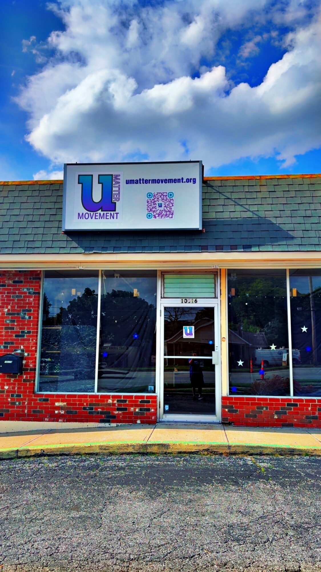 Storefront with a sign displaying a logo and QR code, brick exterior, large front windows, and a door, under a cloudy sky.