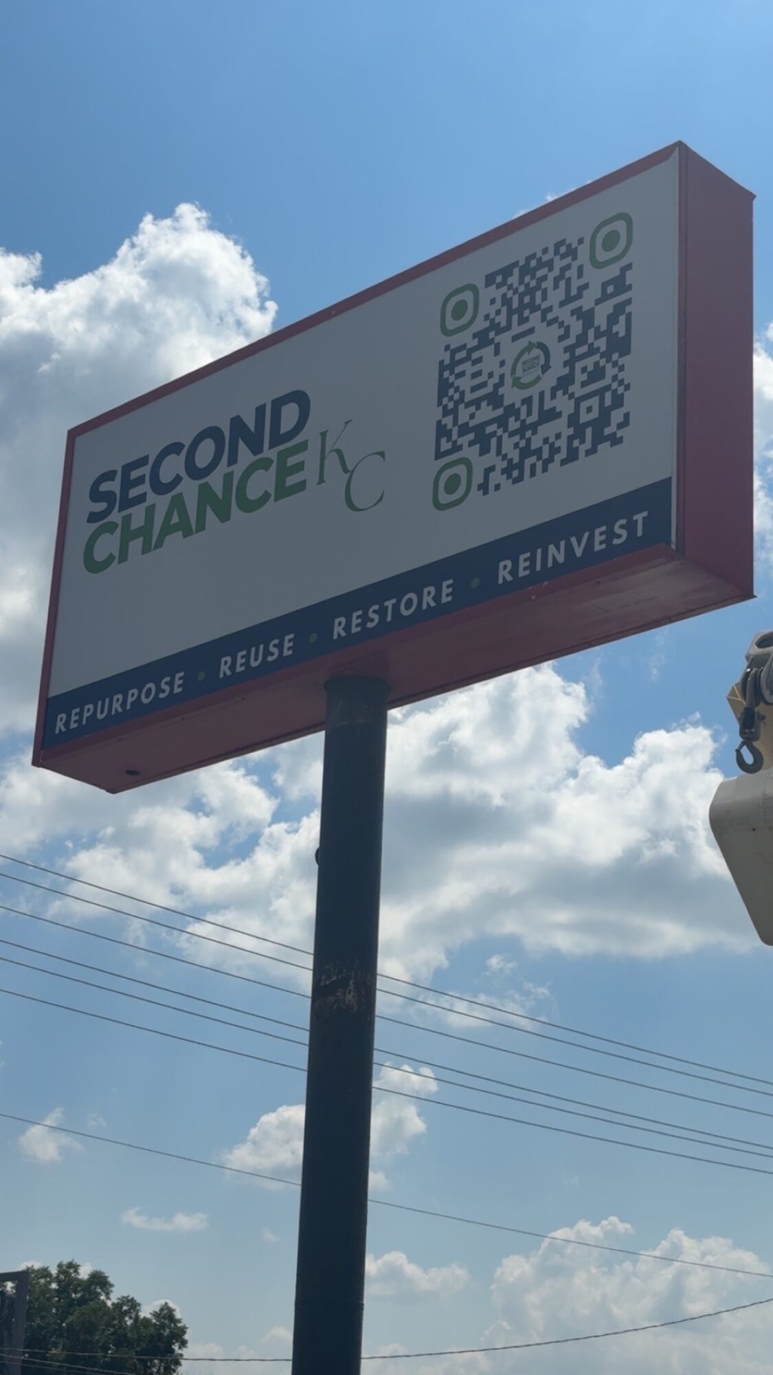 Billboard with QR code and text reading 'SECOND CHANCE' and 'EFFECTIVE, REUSE, RESTORE, REINVEST' against a blue sky with clouds.