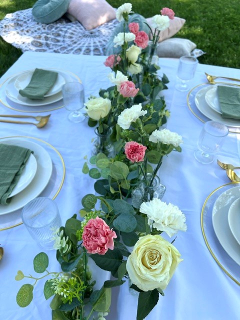 Table with floral centerpiece, plates, glasses, and napkins outdoors on grass, with a blanket and pillows in background.
