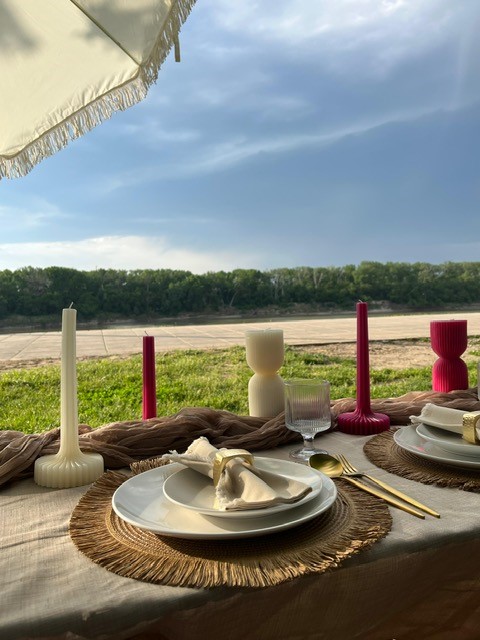 Table set outdoors with plates, candles, and glassware under a canopy, overlooking a grassy field and blue sky.