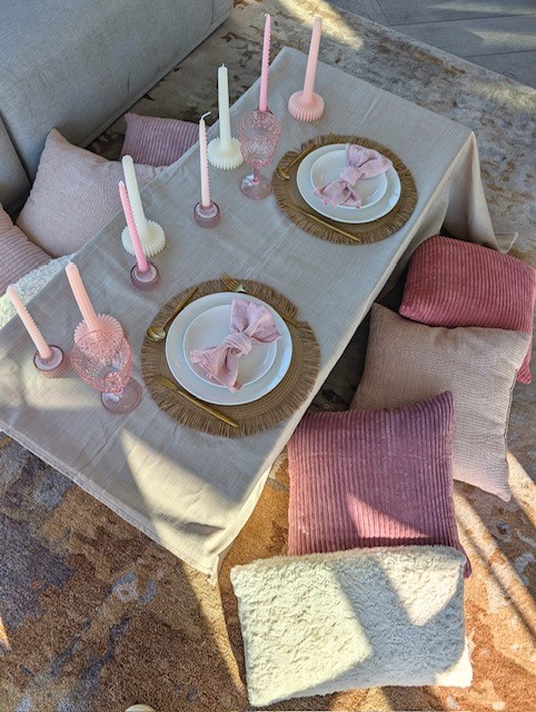 Table with pink candles, plates with pink napkins, surrounded by pink and cream pillows, sunlight casting shadows.