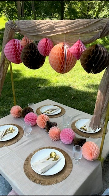 Outdoor table decorated with pink, black, and peach paper lanterns and pom-poms, set with plates, napkins, and glasses.