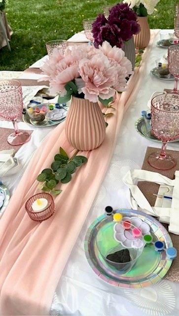 Table decorated with pink flowers in vases, pink table runner, and glassware, set outdoors on grass.