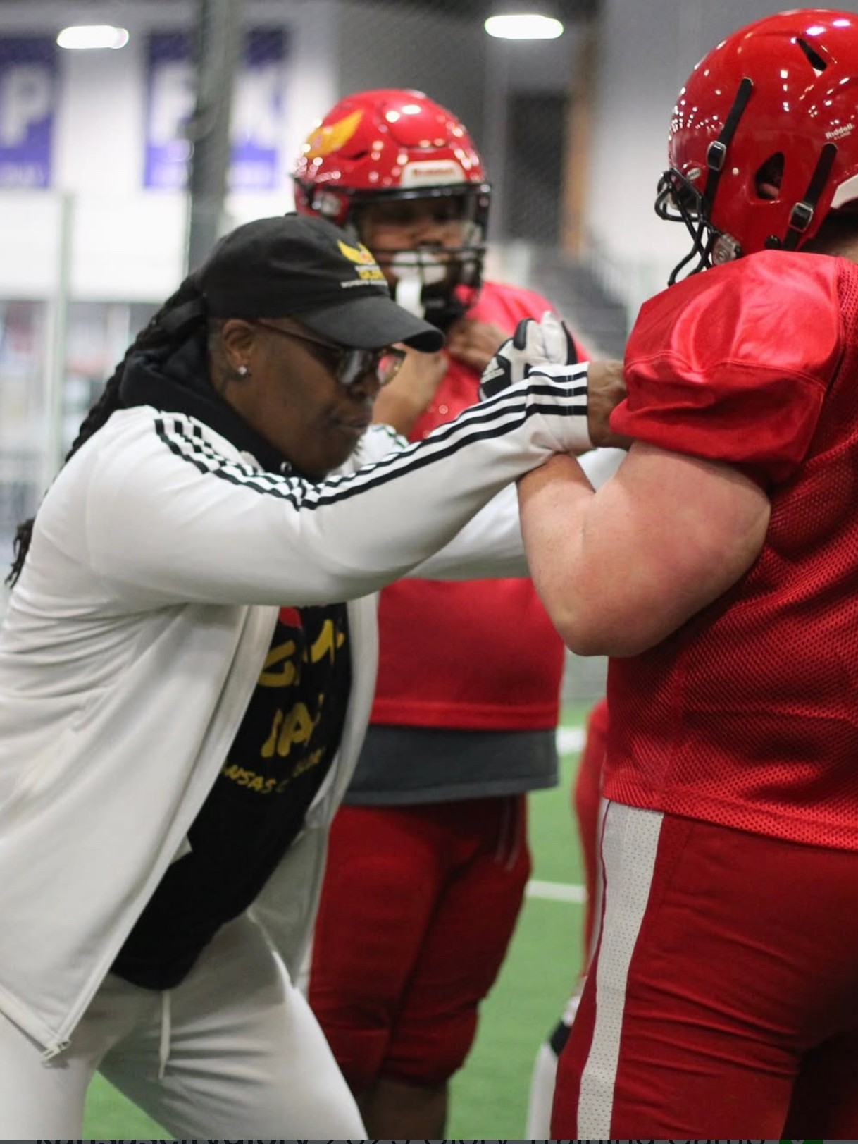 Coach adjusting player's helmet during football practice, with another player in the background, all wearing helmets and sports gear.
